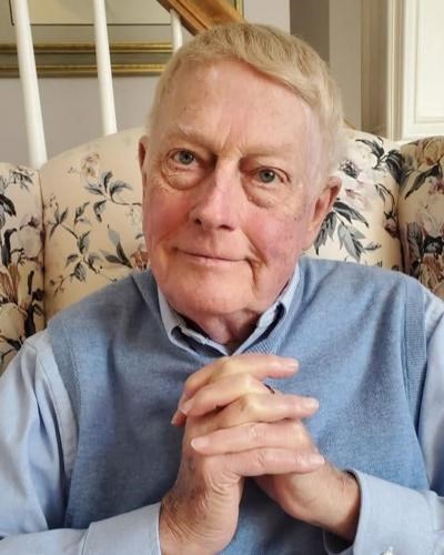Richard Floyd Smith, an elderly man with short, neatly combed hair, sits on a floral-patterned couch wearing a light blue sweater vest over a collared shirt. He smiles warmly at the camera with his hands gently clasped.