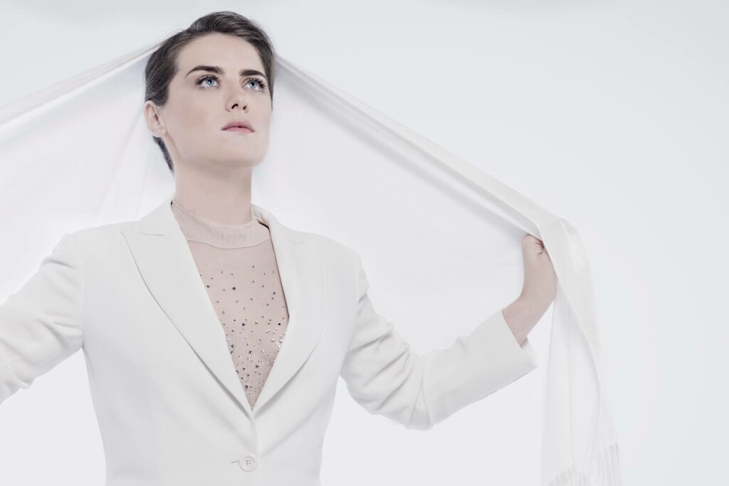 Alessandra Maestrini stands poised in a bright white suit and sheer rhinestone blouse, holding a white scarf over her shoulders against a pure white backdrop. Her expression is serious and upward-facing.