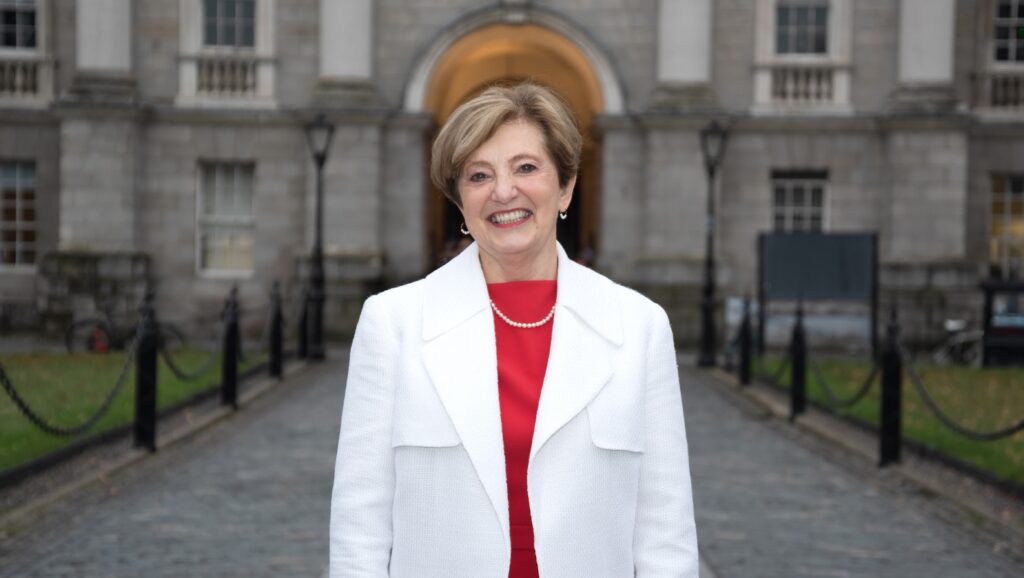 Katherine Zappone in a red dress and white coat standing proudly in front of the entrance to Trinity College Dublin, smiling directly at the camera.