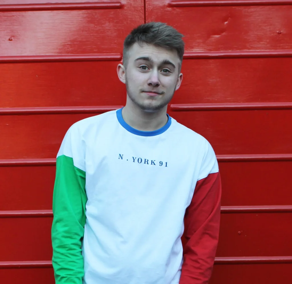 Alex Bertie standing in front of a bright red wall, wearing a white long-sleeved shirt with green and red sleeves and the words "N. YORK 91" across the front. He has short, styled hair and a soft smile, looking directly at the camera.