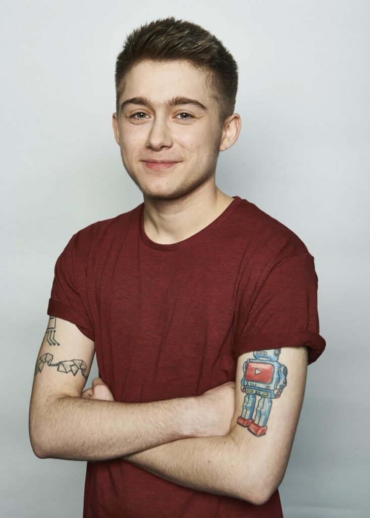 Alex Bertie wearing a maroon t-shirt, smiling with arms crossed. Tattoos are visible on both arms, including a colorful robot with a YouTube play button. He stands against a light gray backdrop.