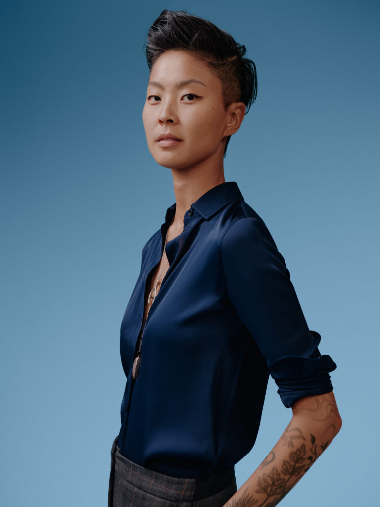 risten Kish posing confidently in a dark blue silky blouse and tailored gray trousers, with her arms crossed and tattoos peeking out on a blue background.