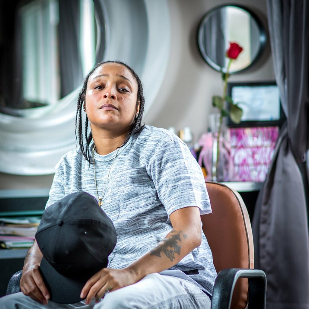 Felicia Pearson seated indoors in a casual gray T-shirt and white pants, holding a black cap in one hand. A red rose in a vase is visible in the softly focused background. She gazes forward with a relaxed demeanor.