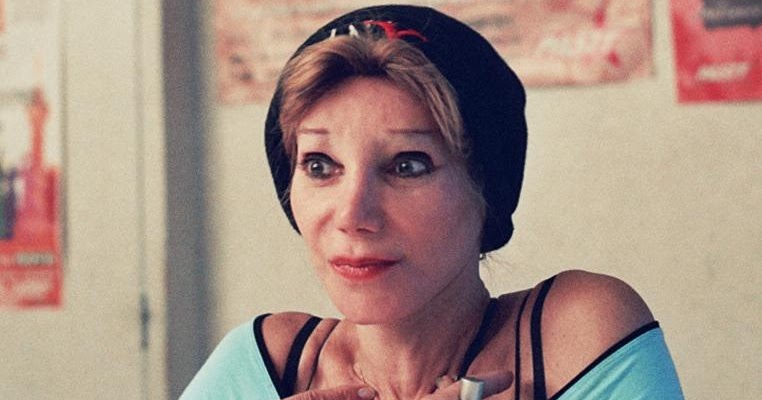 A younger Camille Cabral wearing a black beanie and a light blue top, looking alert and expressive with her hands placed over her chest.