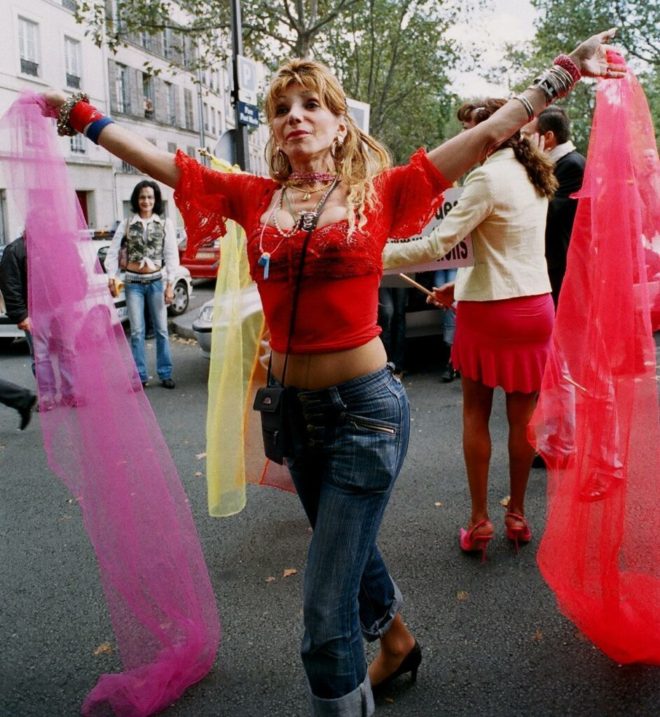 Camille Cabral dancing in the street during a protest, wearing a red lace top, blue jeans, and colorful scarves, with arms extended outward joyfully.