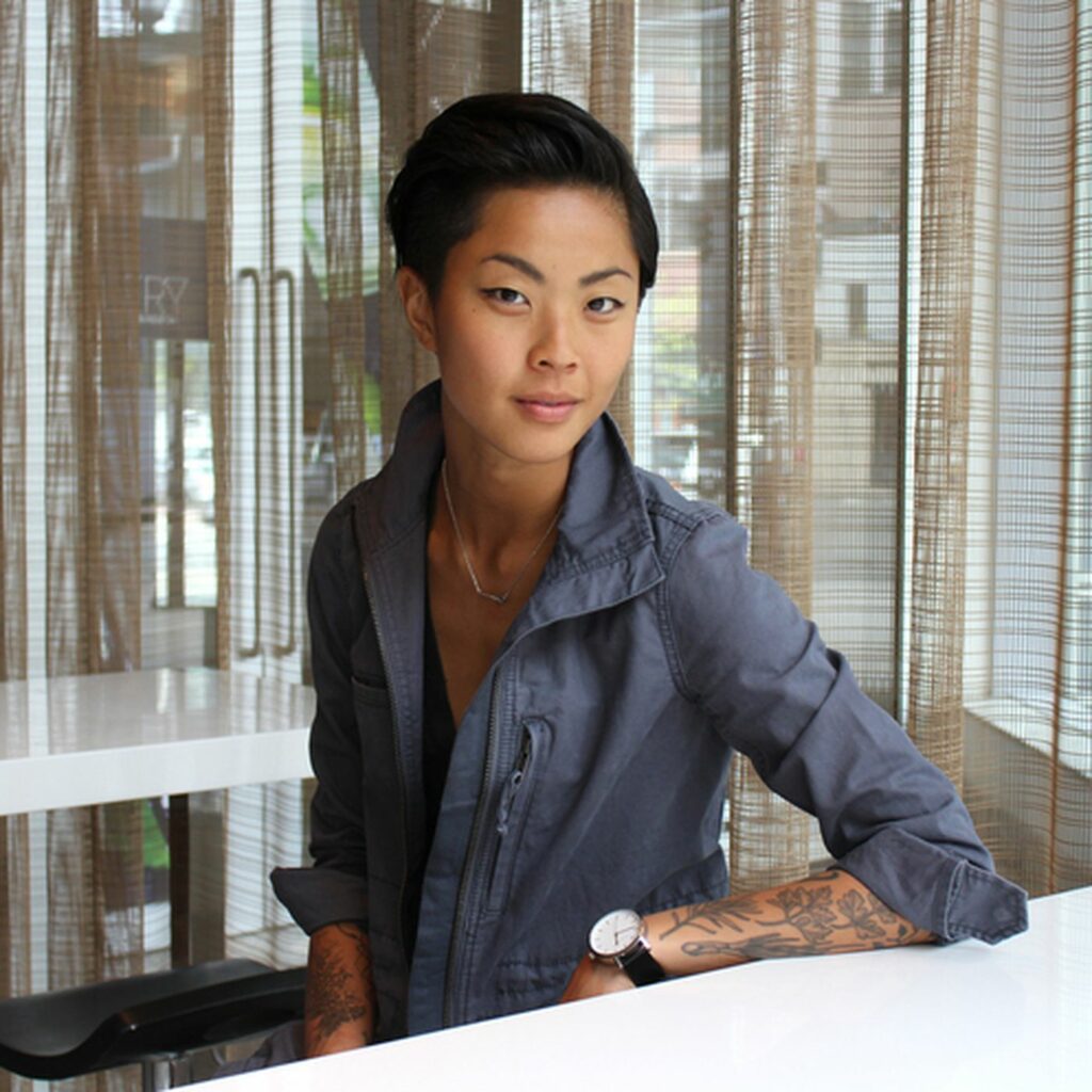 Kristen Kish seated at a white counter in front of sheer window blinds, wearing a dark gray zippered jacket and a wristwatch, with a relaxed expression.