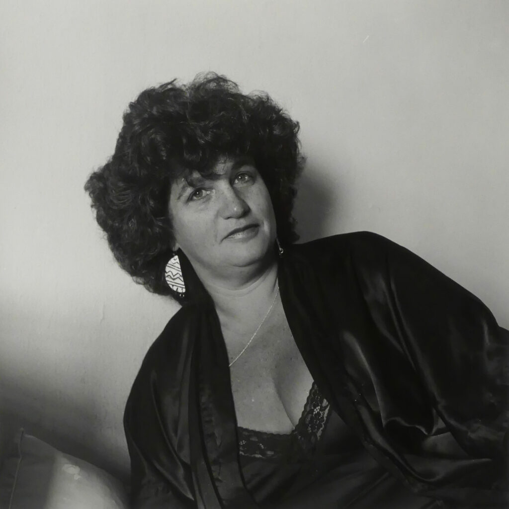 Black and white portrait of Joan Nestle reclining in a dark satin robe, wearing large patterned earrings and a lace-trimmed camisole, with a soft expression.