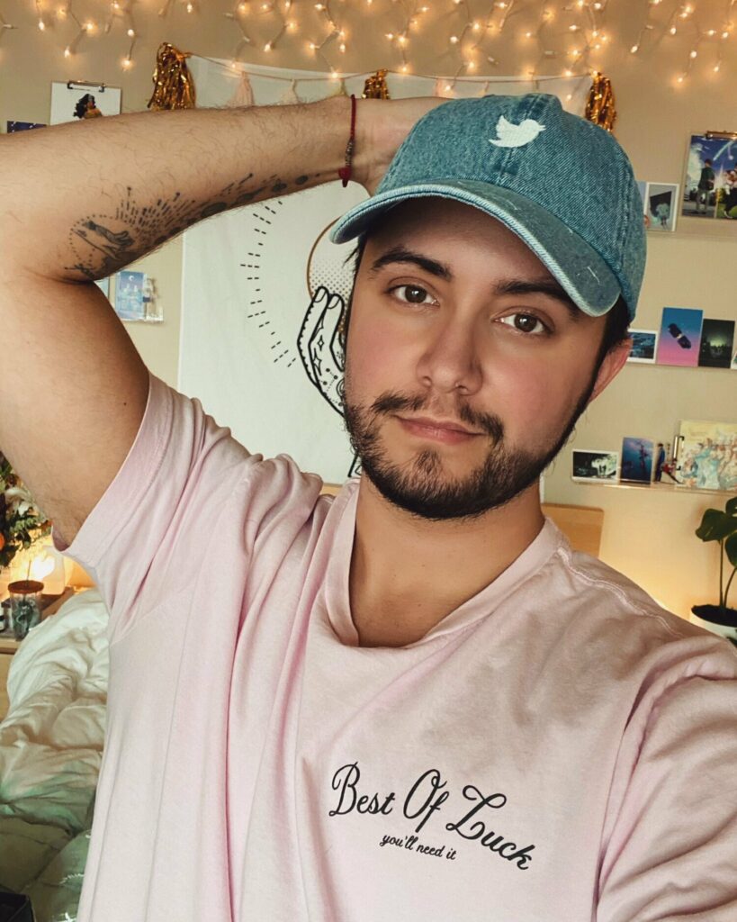 Aiden Thomas poses in a pink shirt reading “Best of Luck, you’ll need it,” with one arm behind his head. He wears a denim Twitter cap and stands in a cozy, decorated room with string lights and art.