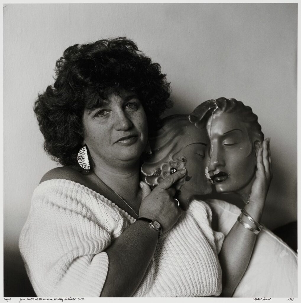 Joan Nestle in a white off-the-shoulder sweater holding a ceramic sculpture of two women’s faces in a kiss, looking directly at the camera in a 1980s black and white portrait.