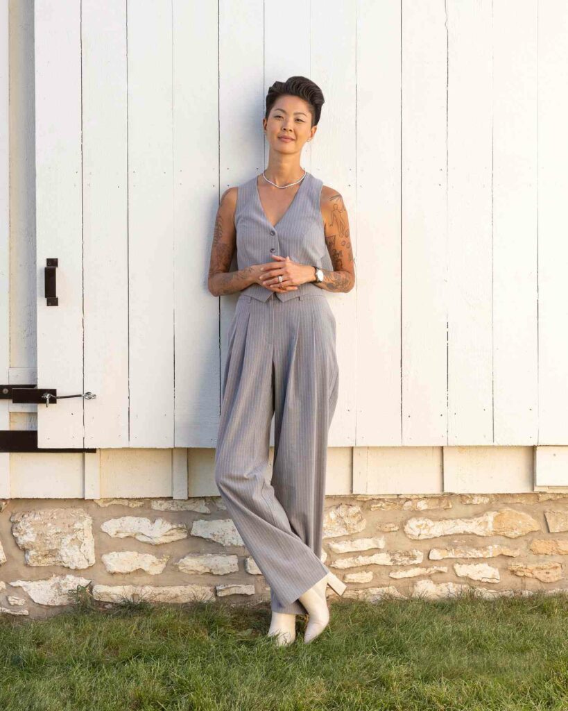 Kristen Kish standing outside against a white wooden wall, wearing a light gray pinstriped vest and wide-leg pants, with tattoos visible on both arms and hands clasped.