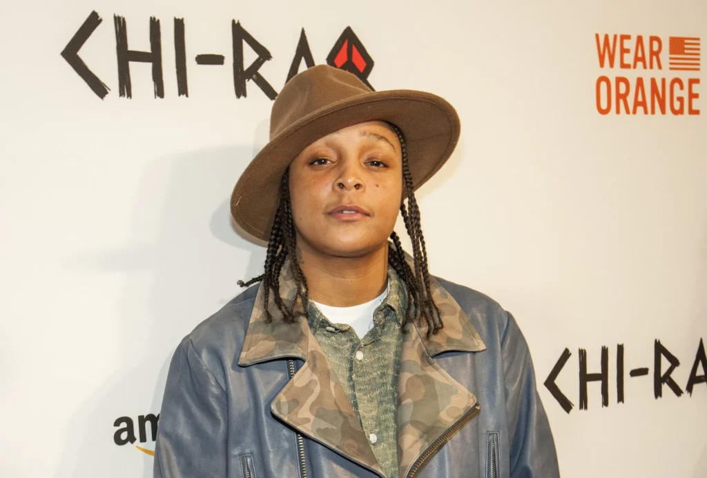 Felicia Pearson posing at a red carpet event for Spike Lee’s Chi-Raq. She wears a brown wide-brim hat, camo-collared shirt, and a blue leather jacket, standing against a white backdrop with “Wear Orange” and “Chi-Raq” logos.