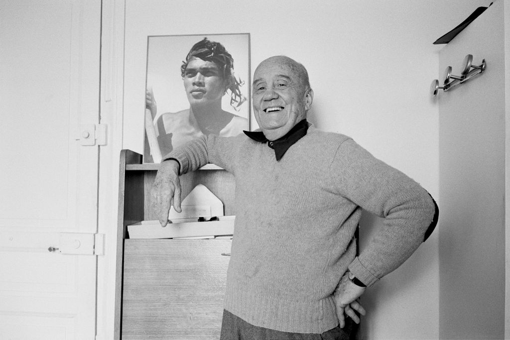 Daniel Guérin in his later years, smiling and leaning casually against a piece of furniture, with a large photograph of a shirtless young man in the background.