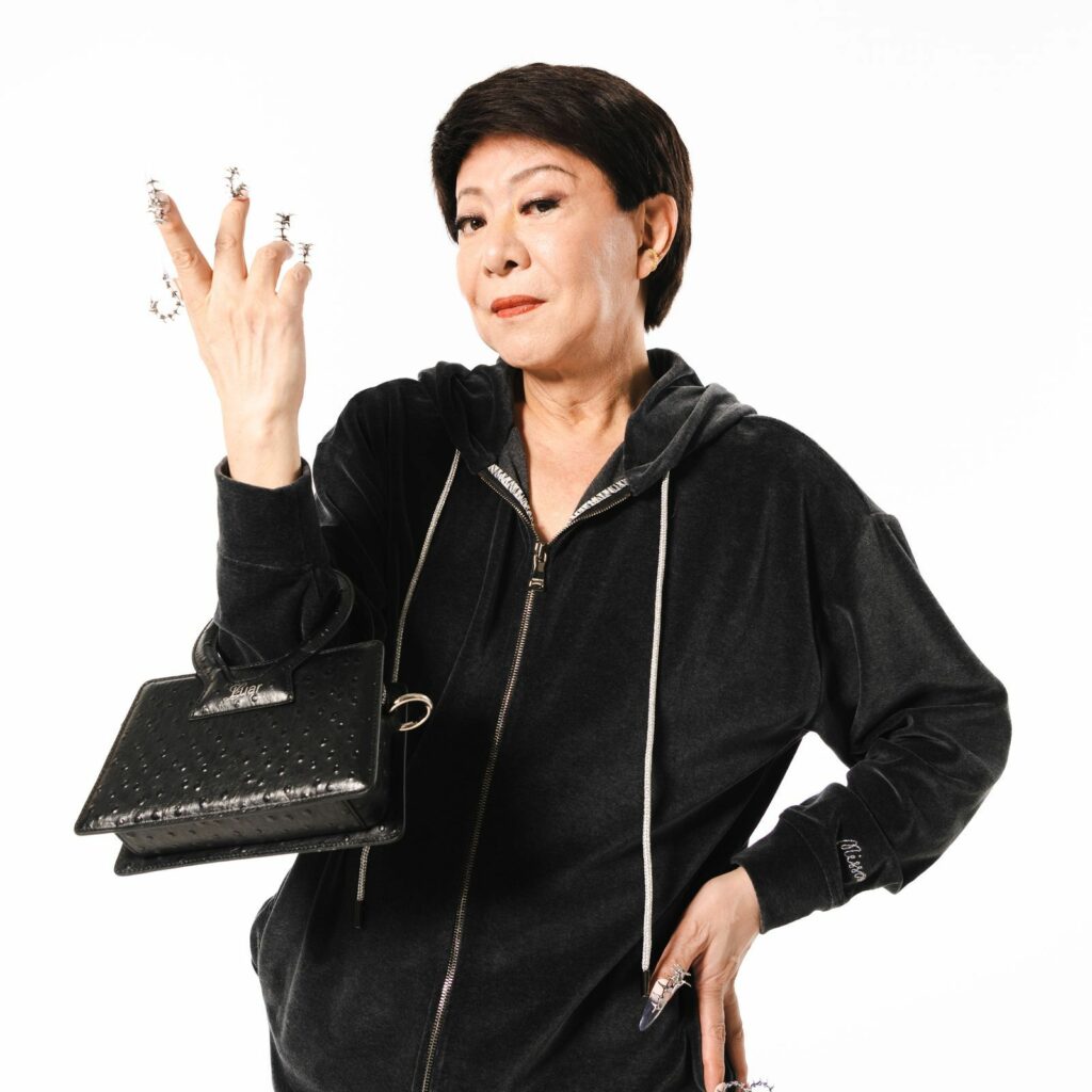 Kenichi Mikawa poses confidently in a black velvet zip-up tracksuit, holding a small black handbag with one hand and flaunting elaborate decorative nails with the other. His expression is cool and composed, exuding flair and attitude.