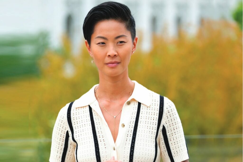 Kristen Kish outdoors in a cream knit polo shirt with black stripes, wearing a subtle necklace and earrings, standing in front of blurred autumn foliage.