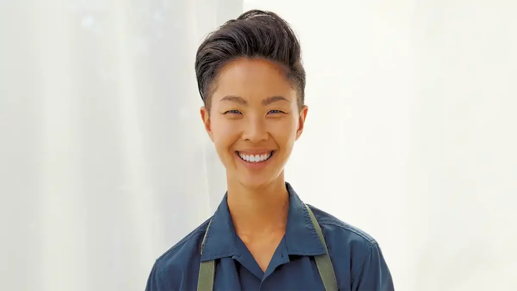 Kristen Kish smiling brightly in a navy blue collared shirt with apron straps over her shoulders, standing against a softly lit white background.