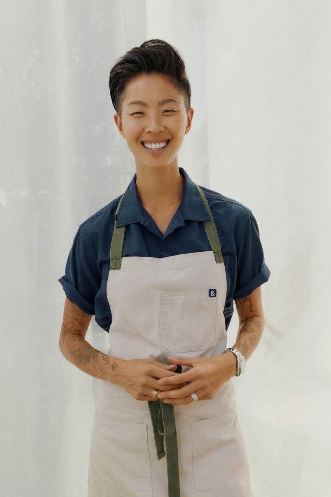 Kristen Kish smiling warmly in front of a white curtain, wearing a navy shirt and beige apron with green straps, hands gently holding the apron ties.