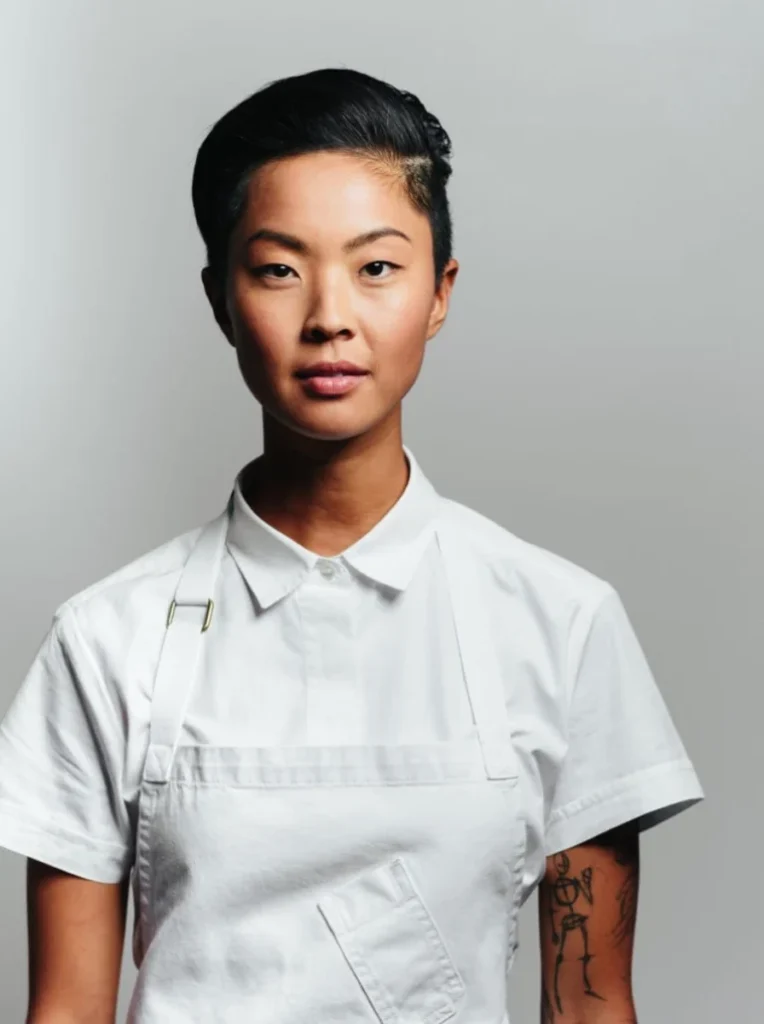 Kristen Kish looking directly at the camera with a composed expression, wearing a white chef's coat and apron against a pale gray background.