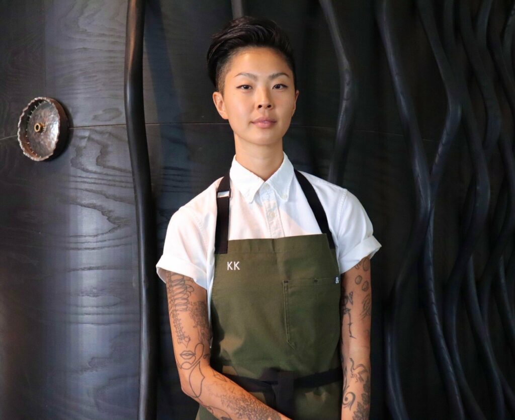 Kristen Kish standing confidently in front of a dark textured wall, wearing a white shirt and olive apron with “KK” embroidered, showcasing her full-sleeve tattoos.