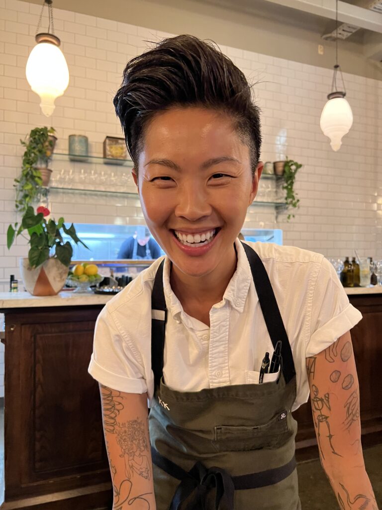 Kristen Kish smiling widely behind the counter at her restaurant Arlo Grey, wearing a white short-sleeve shirt under a gray apron, with tattoos and pens in her apron pocket.