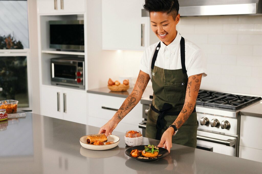 Kristen Kish arranging two plated dishes on a kitchen counter, smiling down at the food while wearing a white shirt, dark apron, and surrounded by kitchen tools and a jar of kimchi.