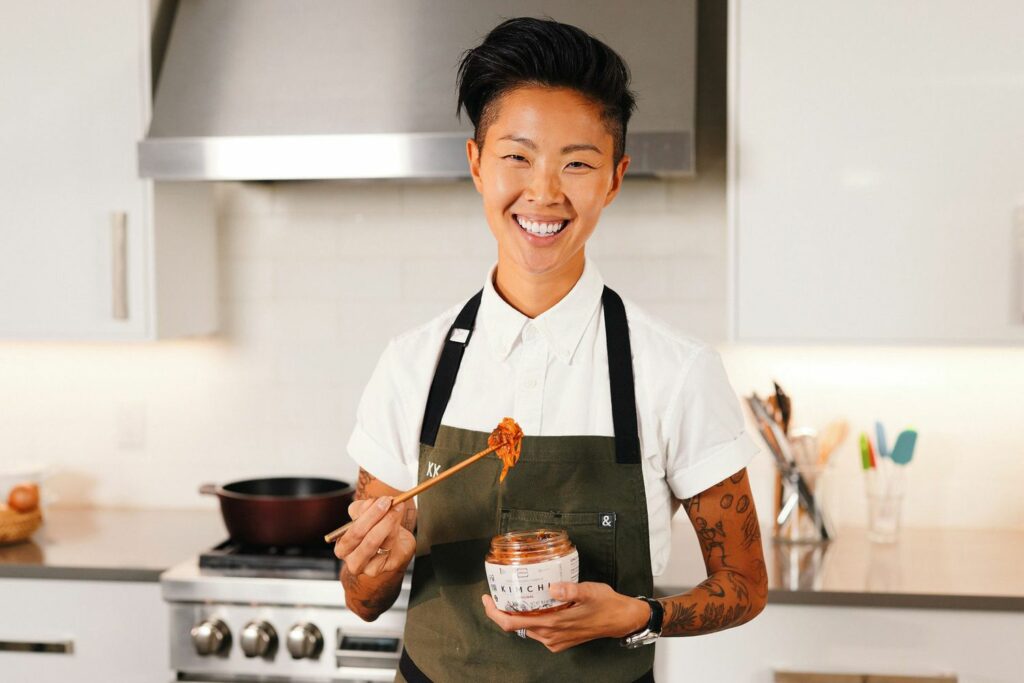 Kristen Kish smiling in a modern kitchen, holding a jar of kimchi in one hand and a chopstick with a bite of it in the other, wearing a white shirt and olive apron embroidered with “KK.”
