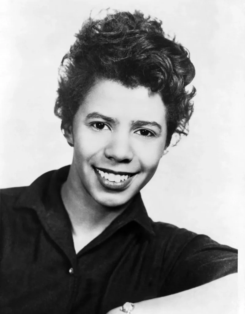Studio portrait of a young Lorraine Hansberry in a collared shirt, smiling widely with a clean white backdrop.