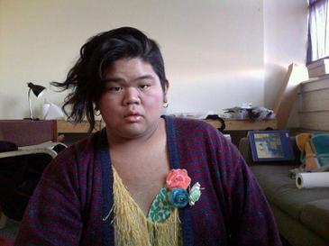 CallOutQueen sits in a lived-in studio space, wearing a cozy cardigan over a fringed yellow top adorned with colorful fabric flowers. Her hair is styled in waves swept to one side, and she meets the camera with a steady, thoughtful look.