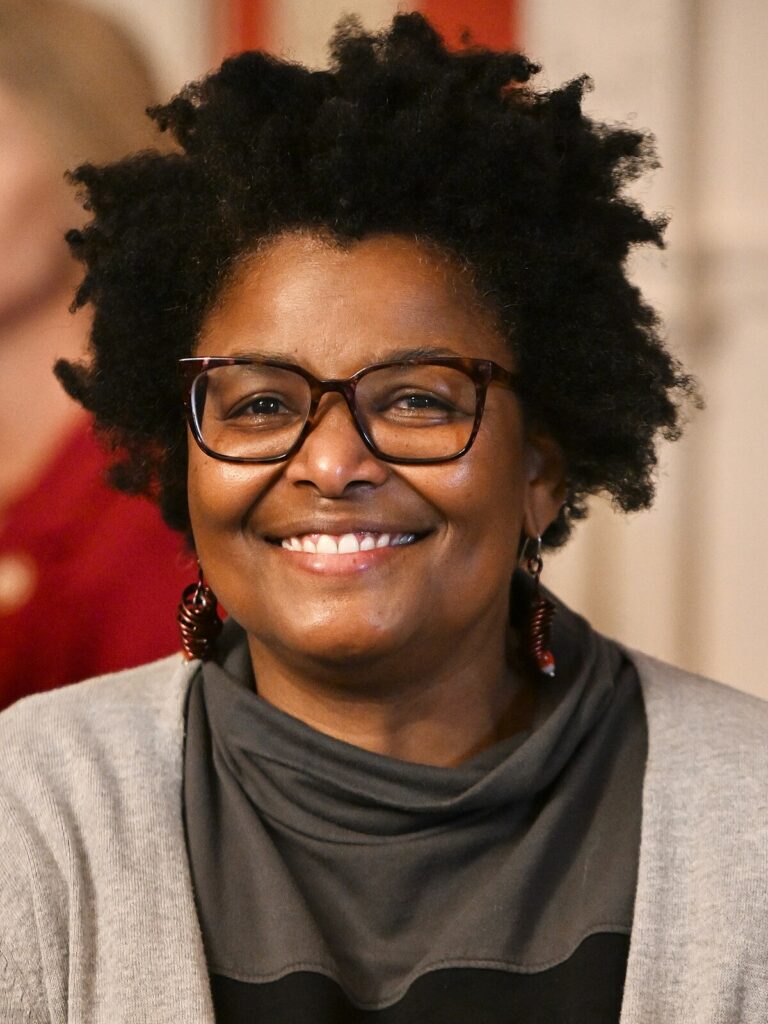 Mary L. Washington smiling during an event, wearing a gray and black top with large earrings and brown glasses.