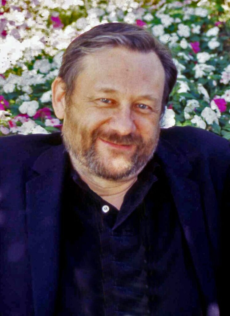 A smiling Michael Bronski seated outdoors in front of a vibrant floral background. He wears a dark blazer and has a full beard, giving a warm and approachable look.
