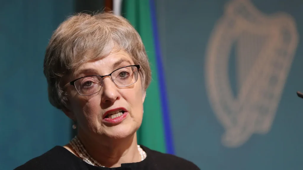 Katherine Zappone speaking at a podium with a serious expression, framed by an Irish government harp symbol and flag in the background.
