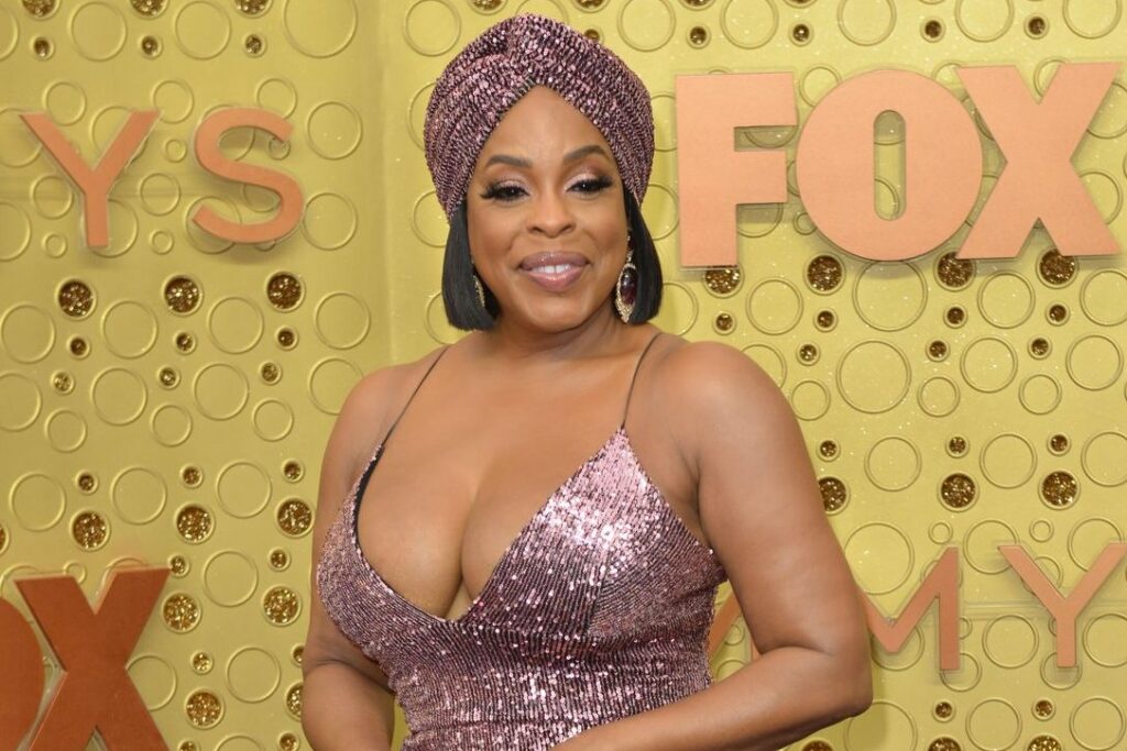 Niecy Nash-Betts poses in front of a gold Emmy Awards backdrop wearing a sparkling rose-pink dress and a matching turban, smiling warmly with bold lashes and statement earrings.