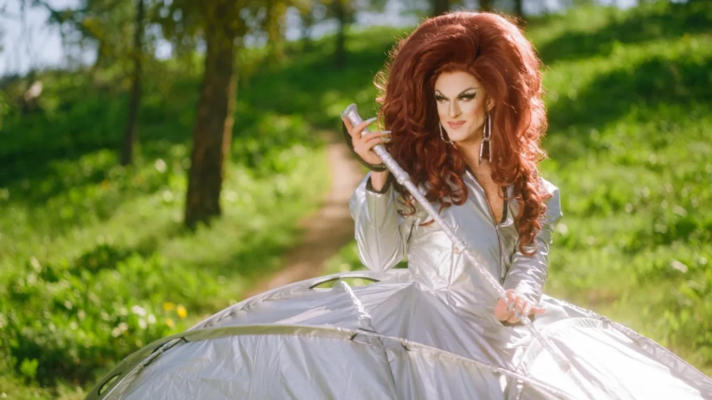 Pattie Gonia smiles while holding a silver trail counter device and wearing a large structured metallic silver gown. She stands on a green wooded trail surrounded by sunlight and trees.