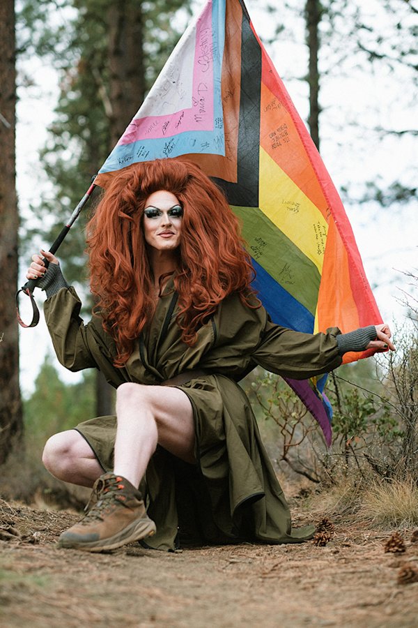 Pattie Gonia kneels in a forest holding a Progress Pride flag over her shoulder, wearing a loose olive trench dress, full makeup, voluminous red hair, and hiking boots.

