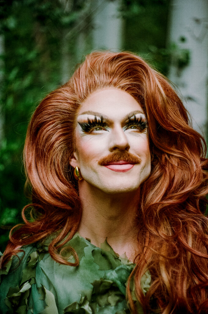 Pattie Gonia gazes upward with a peaceful smile while wearing dramatic makeup, a leafy green outfit, and voluminous red hair in a forest setting.