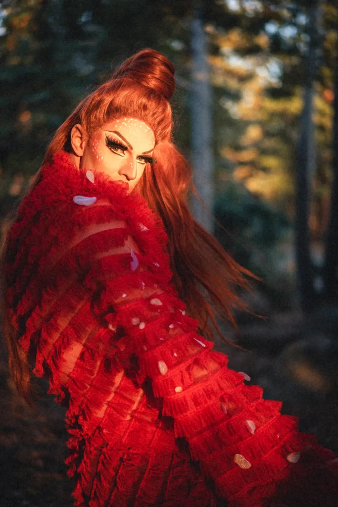 Pattie Gonia turns toward the camera with intensity, bathed in golden light. She wears a sheer red ruffled garment, bold white makeup dots, and high-volume red hair in a pine forest.