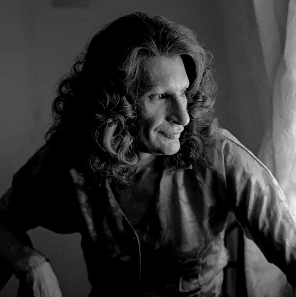 A monochrome close-up of Sylvia Rivera seated indoors by a curtain, her long hair loose and her gaze focused off-camera, capturing a contemplative and powerful mood.