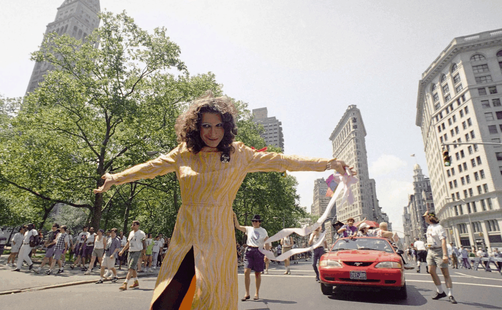 A vibrant color photo of Sylvia Rivera in a yellow striped dress during a Pride parade, arms extended joyfully in the street as crowds march behind her near the Flatiron Building in New York City.