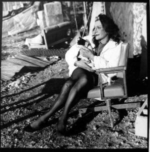 A candid black-and-white photograph of Sylvia Rivera sitting in a chair outdoors, dressed in black tights and a light top, holding a black-and-white cat in her arms with tenderness.