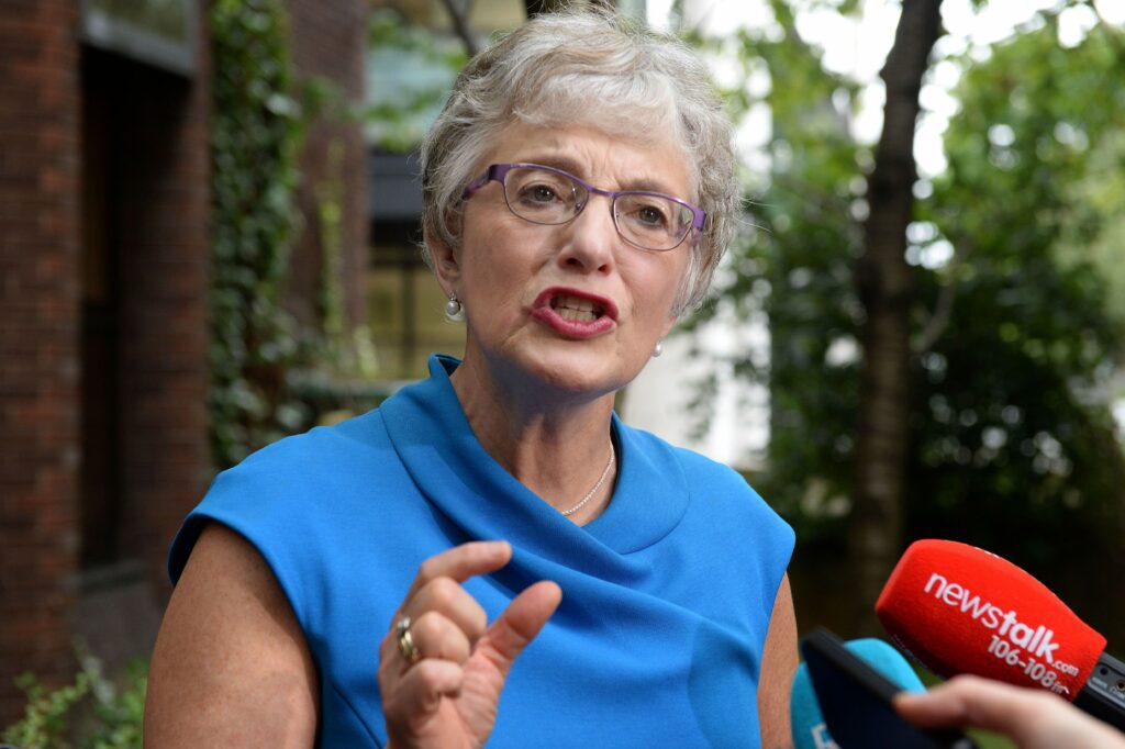 Katherine Zappone addressing reporters while wearing a bright blue dress, gesturing with her hand, and speaking into red and teal NewsTalk microphones.