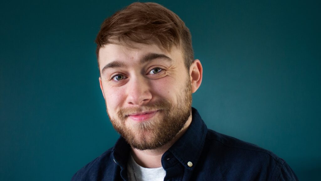 Close-up of Alex Bertie with a full beard and styled hair, smiling softly while wearing a dark blue button-down shirt. The background is a solid teal color, giving the image a calm and professional tone.