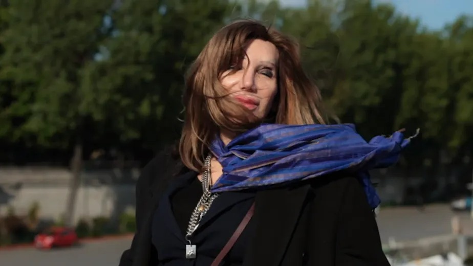 Camille Cabral outdoors with windblown hair, wrapped in a flowing blue scarf, dressed in black, standing near a river with trees in the background.