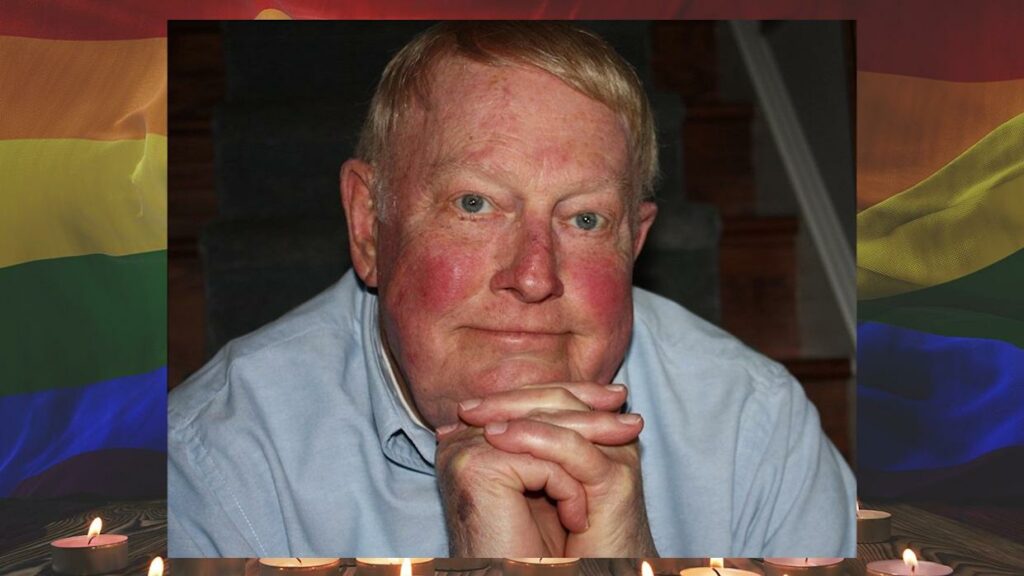 Richard Floyd Smith rests his chin on his folded hands in a solemn memorial portrait. The background features a rainbow pride flag and glowing candles, honoring his legacy as a beloved gay educator.