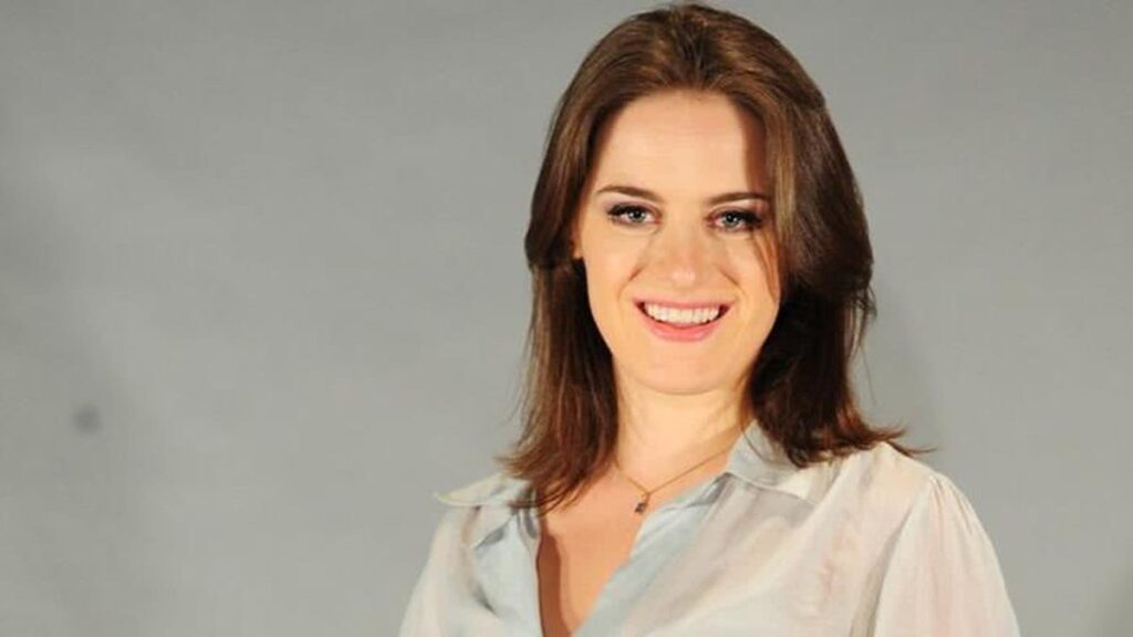 Alessandra Maestrini smiles brightly in a light blouse, hair down, standing against a neutral gray background.