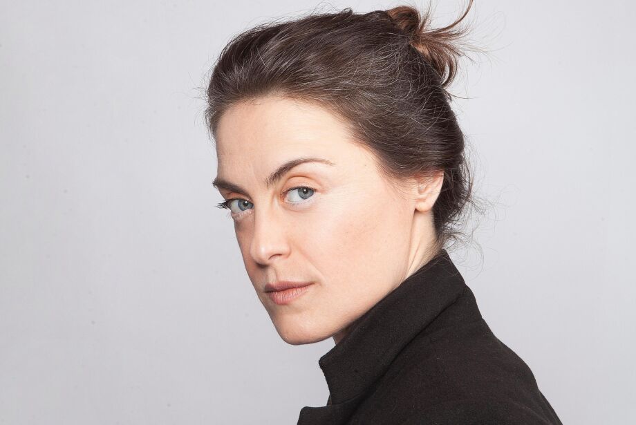 Alessandra Maestrini poses in a black coat with her hair tied up, gazing over her shoulder with a confident, serious expression against a light gray background.