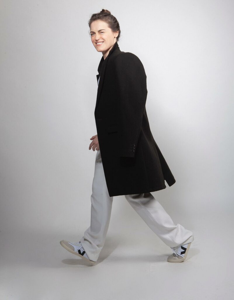 Alessandra Maestrini is mid-step in white trousers, a black coat, and sneakers, looking playful and smiling as she walks across a light-colored studio set.