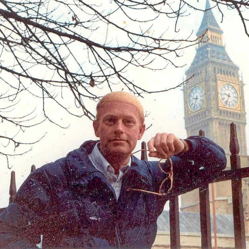 A younger Richard Floyd Smith stands in front of Big Ben in London, holding his glasses and wearing a dark jacket. Bare tree branches surround him in the wintry scene, capturing his years of travel and teaching abroad.