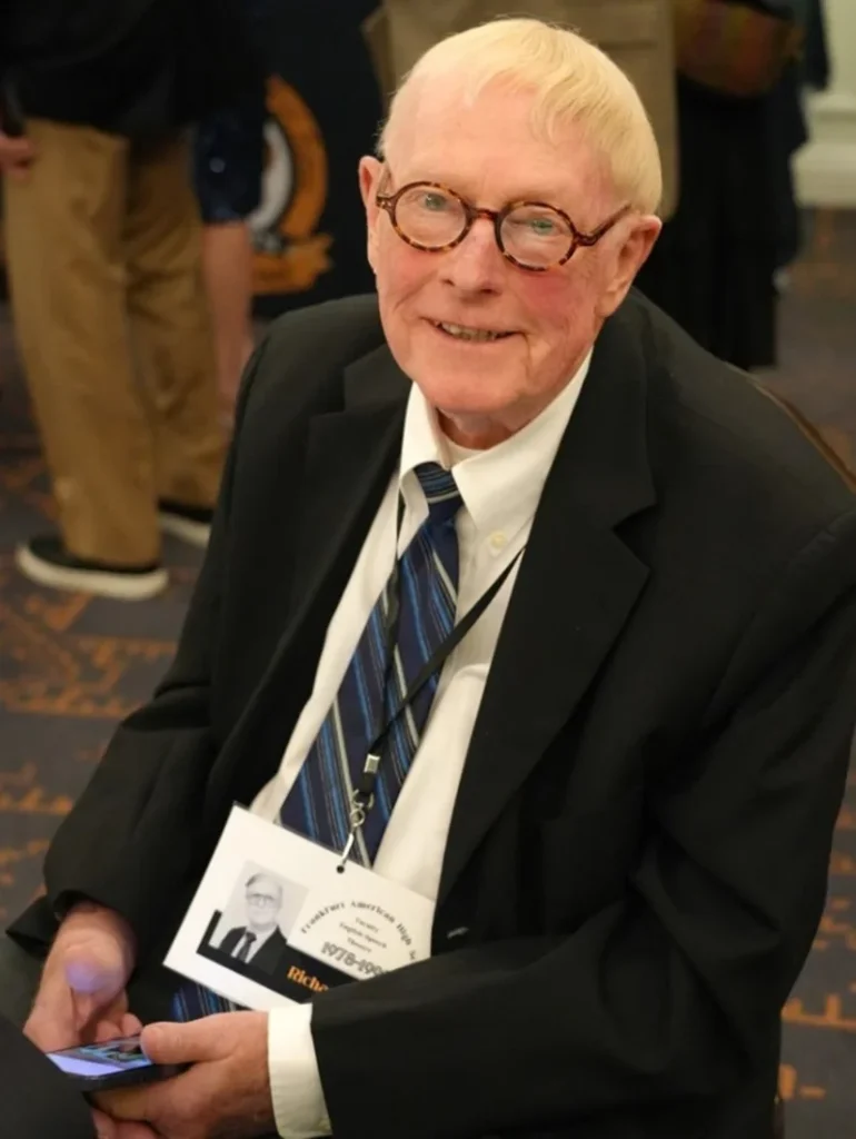 Richard Floyd Smith smiles while seated at a formal event wearing a black suit and striped tie. He holds a phone in his hand, and a name badge with his portrait hangs from his neck.