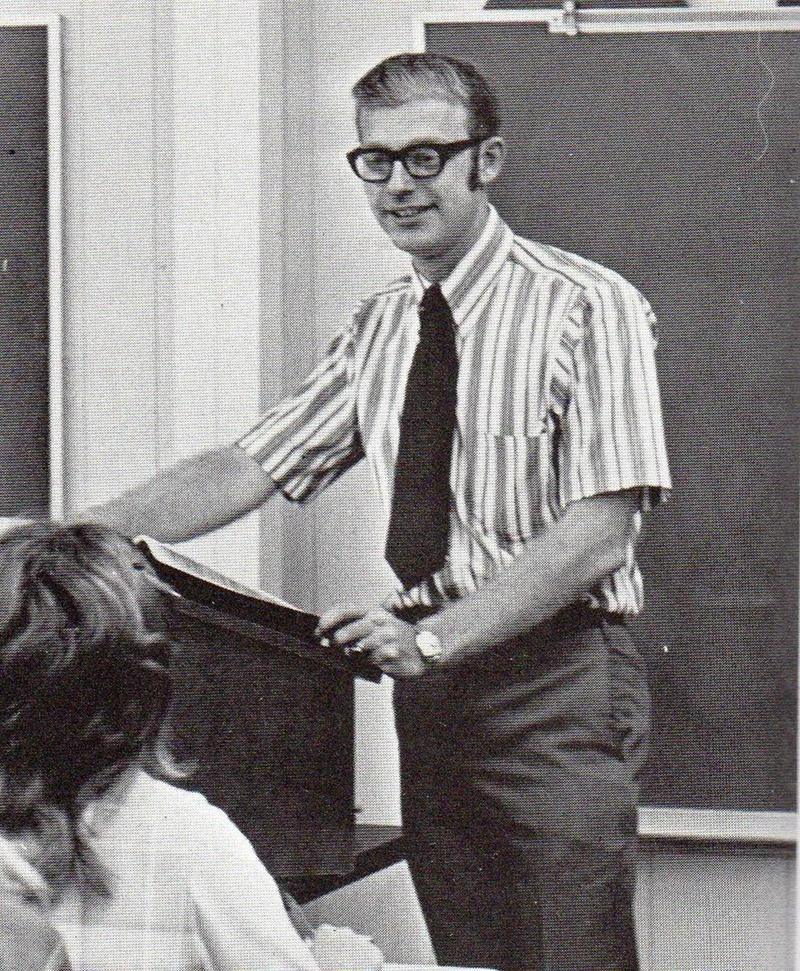 A young Richard Floyd Smith stands at a podium in a 1970s classroom, mid-lecture, wearing a striped shirt and tie with thick-rimmed glasses. He engages his students with confidence and clarity.