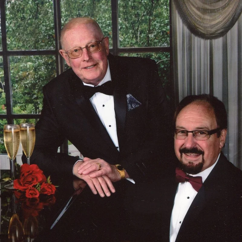 Richard Floyd Smith and his husband Lynn Barnes pose in tuxedos beside a piano adorned with champagne flutes and roses. They smile warmly in a formal studio portrait celebrating their enduring partnership.