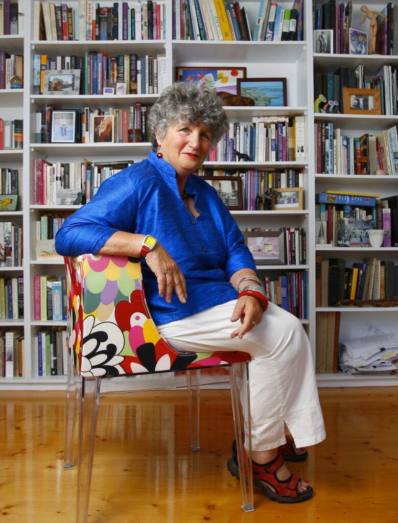 Joan Nestle seated in a colorful floral-patterned chair, wearing a bright blue shirt and white pants, surrounded by bookshelves filled with books and photographs in a warmly lit room.